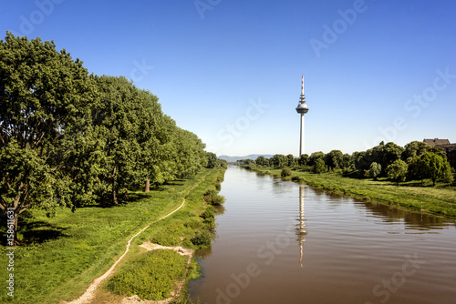 Germany, Baden-Wuerttemberg, Mannheim, Neckar river: Panoramic view of  Telecommunication Tower that  mirrors in the river with green trees, clear blue sky and horizon