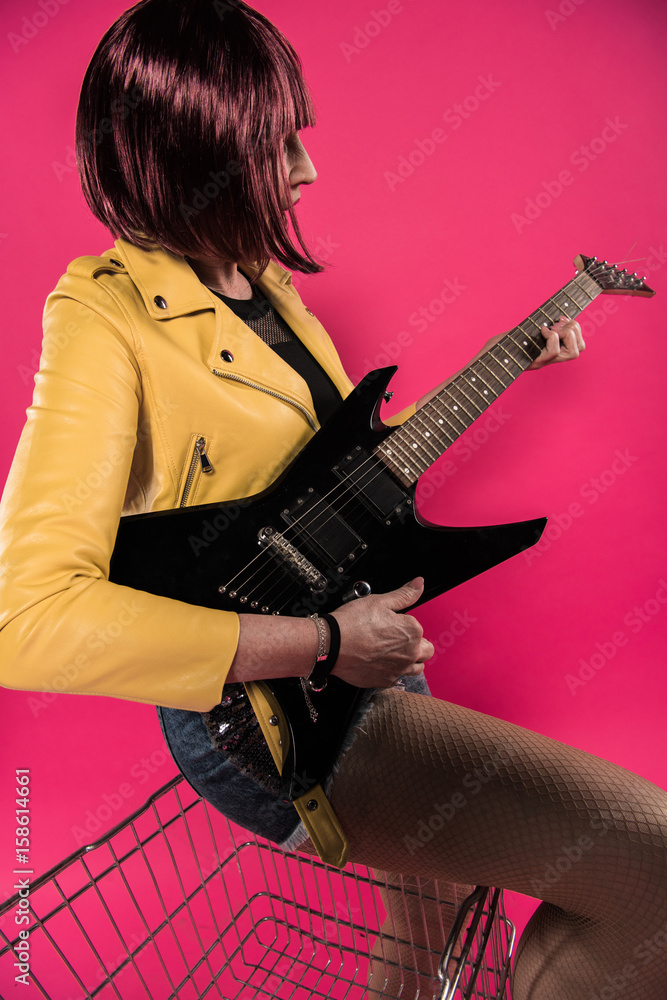 Fototapeta premium Stylish senior woman with pink hair playing electric guitar isolated on pink