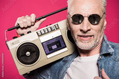 portrait of senior man in sunglasses holding tape recorder isolated on pink