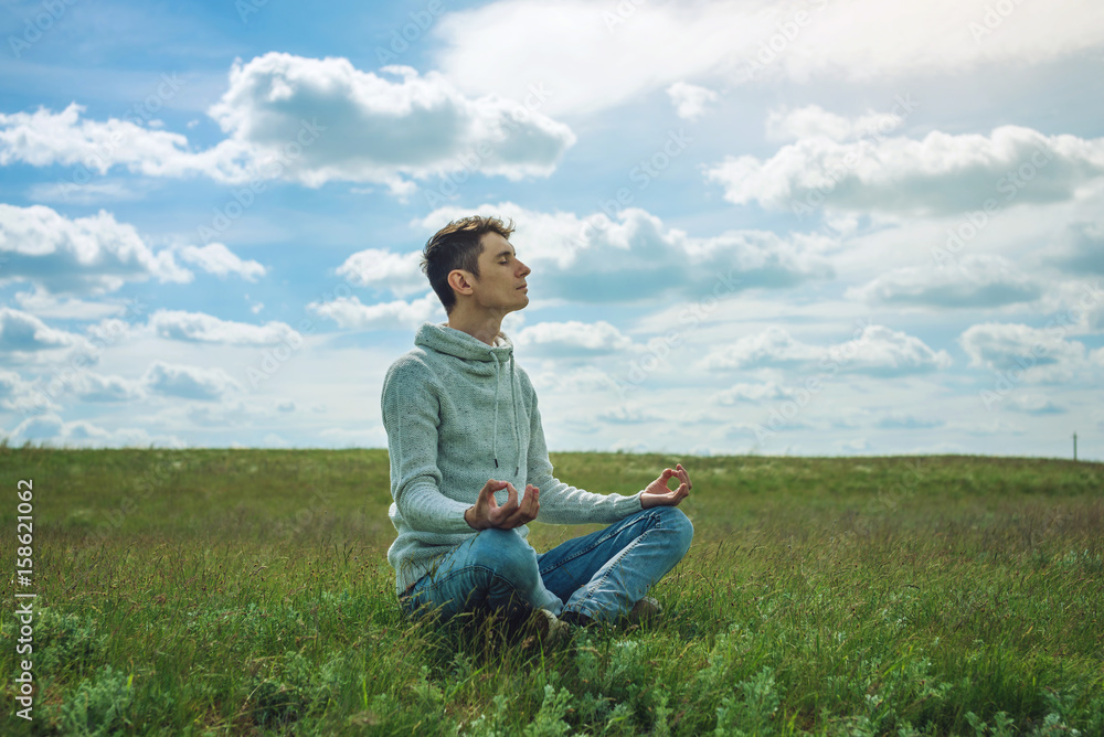 Man traveler with backpack sits on meadow with green grass under blue sky with clouds in the Lotus position Man traveler with backpack sits on meadow with green grass under blue sky with clouds in the Lotus position