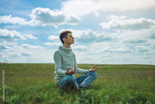 Man traveler with backpack sits on meadow with green grass under blue sky with clouds in the Lotus position © Artem