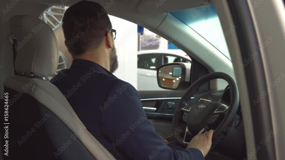 Male caucasian customer showing key inside the car at the dealership. Attractive bearded man turning to the back seat. Close up of handsome guy in glasses moving the sun visor Male caucasian customer showing key inside the car at the dealership. Attractive bearded man turning to the back seat. Close up of handsome guy in glasses moving the sun visor