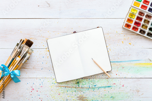 Artistic background design workspace, notebook flat lay mock up for artwork with watercolor paints, pencil and bunch of different paintbrushes on white wooden table. Top viiew from above, copy space photo