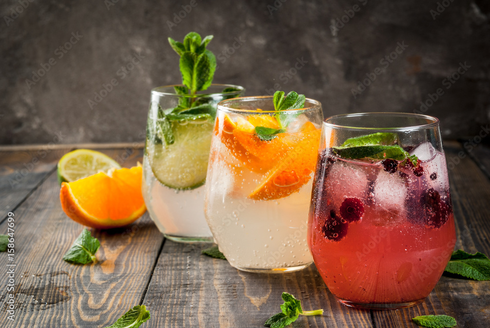 Selection of three kinds of gin tonic: with blackberries, with orange, with lime and mint leaves. In glasses on a rustic wooden background. Copy space Selection of three kinds of gin tonic: with blackberries, with orange, with lime and mint leaves. In glasses on a rustic wooden background. Copy space