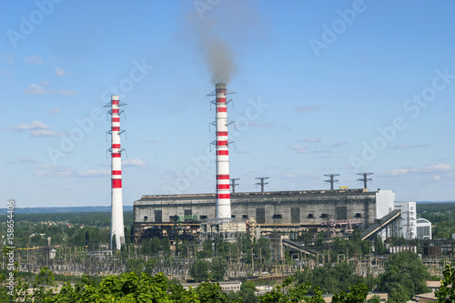 Pipe industrial plant with smoke against bright blue sky and above green trees