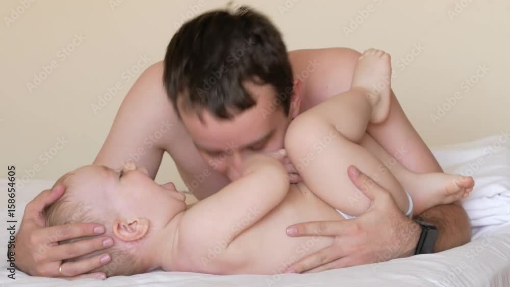 Father kisses and tickles his little son. They lay on the bed after sleeping. The baby is less than two years old. Father kisses and tickles his little son. They lay on the bed after sleeping. The baby is less than two years old.