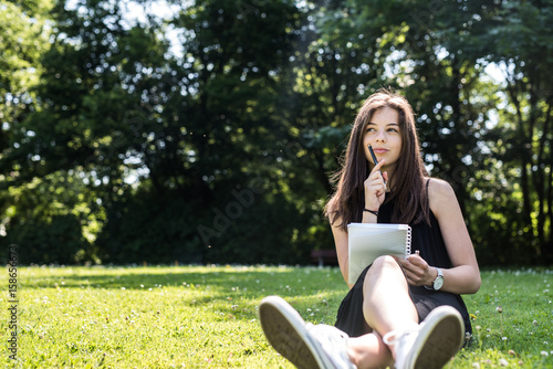 Mädchen sitzt mit Stift und Block auf einer Wiese und überlegt