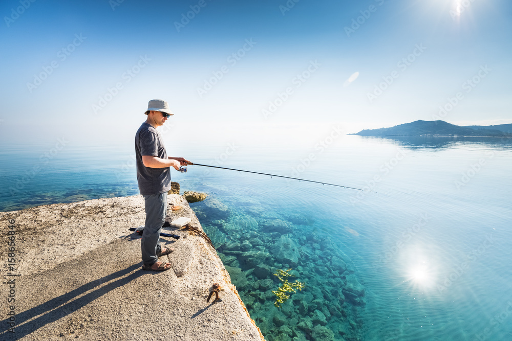 Side view of young man fisherman standing on pier with rod. Seashore of Ionian sea, Zante - Zakinthos island, Greece. Fishing background. Sunrise morning scenery. Side view of young man fisherman standing on pier with rod. Seashore of Ionian sea, Zante - Zakinthos island, Greece. Fishing background. Sunrise morning scenery.