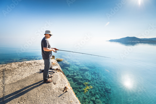 Side view of young man fisherman standing on pier with rod. Seashore of Ionian sea, Zante - Zakinthos island, Greece. Fishing background. Sunrise morning scenery.