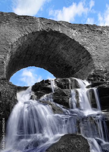 Arched stone bridge with a waterfall and blue sky. Impressive old building with beautiful detail of running water.
