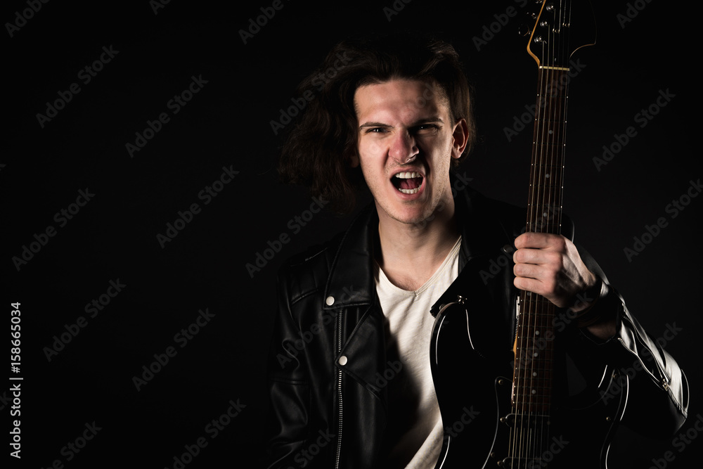 Obraz premium Music and creativity. Handsome young man in a T-shirt, jacket and jeans, with an electric guitar, on a black isolated background. Horizontal frame