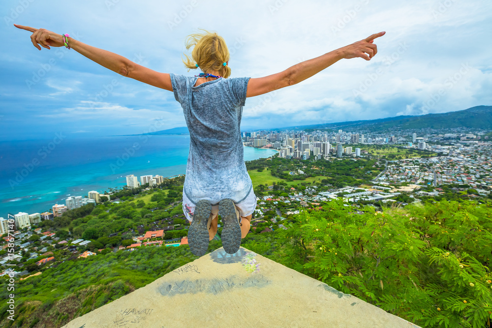 Foto Stock Happy hiker jumping. Hawaiian hiking by popular Diamond Head hike.  Traveler freedom woman. Honolulu and Waikiki Beach Skyline from Diamond Head  State Monument and Park. Oahu Island in Hawaii, USA.