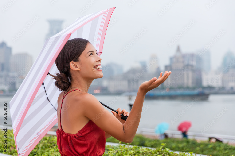 Naklejka premium Deszcz, koncepcja pogody - szczęśliwa azjatycka kobieta turystycznych pod parasolem trzymając rękę do góry sprawdzanie ilości kropel deszczu podczas spaceru po mieście na nabrzeżu w Szanghaju. Młody chiński bizneswoman.