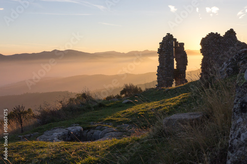 Ruines puit pierres château Pierre Gourde Ardèche avec brume brouillard rayons de lumière soleil couchant dans la vallée et ciel bleu montagnes au fond  © Reddragonfly