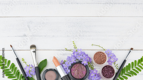 Purple flowers, green leaves, cosmetics and make up brushes on white wood background with copy space
