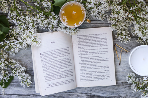 The book is in English surrounded by branches and flowers of white lilac, a mug of tea with lilac petals on a aged wooden vintage background and a white candle in a glass and matches.