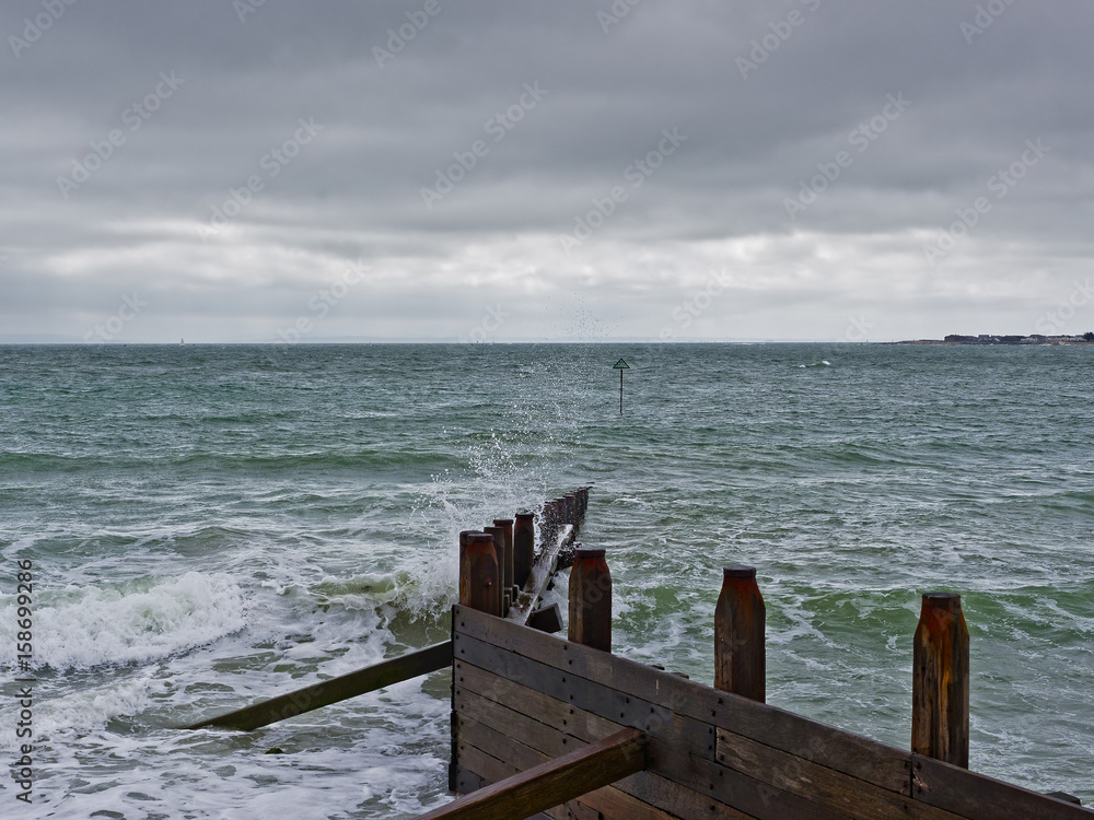 West Wittering beach on a windy day, under a grey sky, waves crash against a partially submerged timber groynes. West Wittering beach on a windy day, under a grey sky, waves crash against a partially submerged timber groynes.