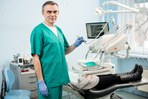 Portrait of handsome male dentist with dental tools in the dental clinic. Doctor wearing green uniform and blue gloves. On the background patient and monitor with X-ray the patient's teeth. Dentistry
