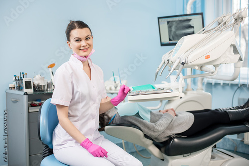 Portrait of friendly female dentist with patient in the dental office. Doctor wearing mask  white uniform and pink gloves. On the background screen with X-ray the patient s teeth