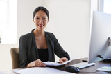 Young Asian woman at desk holding documents smiles to camera