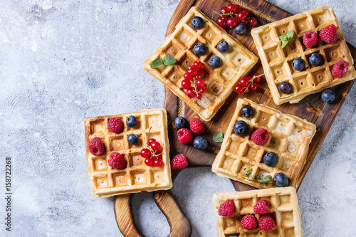 Homemade square belgian waffles with fresh ripe berries blueberry, raspberry, red currant on wooden serving board over gray texture background. Top view with space