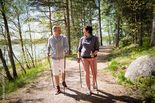 Summer sport in Finland - nordic walking. Man and mature woman hiking in green sunny forest. Active people outdoors. Scenic peaceful Finnish summer landscape.