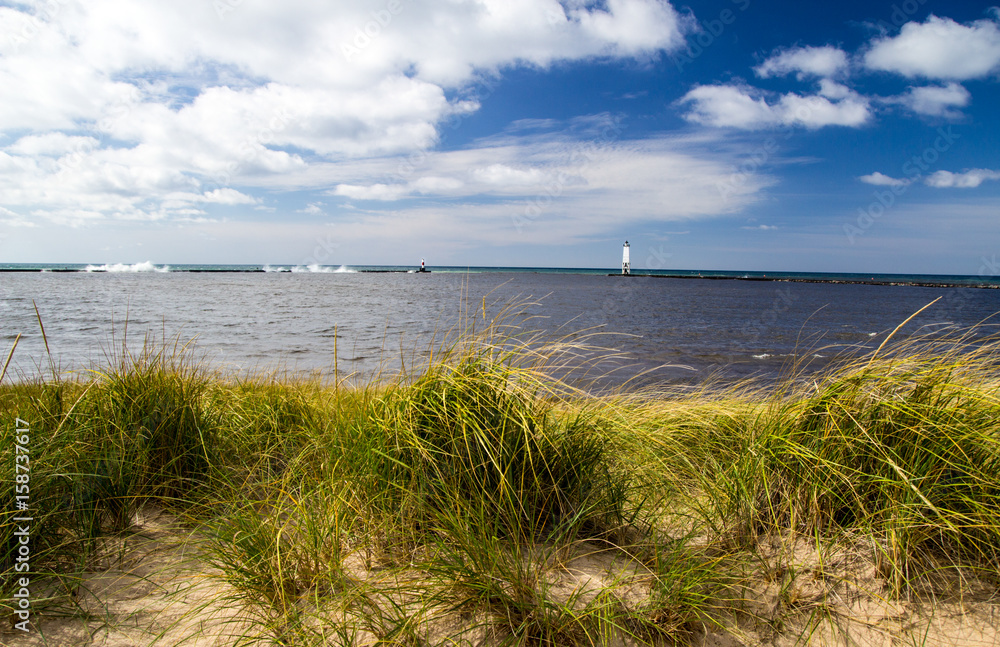 Summer Beach Panorama. Dune grass on the beach with sunny blue sky and lighthouse in the background on the Lake Michigan coast. Frankfort, Michigan. Summer Beach Panorama. Dune grass on the beach with sunny blue sky and lighthouse in the background on the Lake Michigan coast. Frankfort, Michigan.
