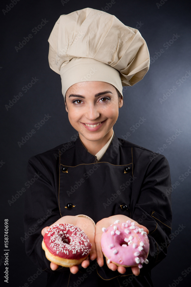 Chef donna con cappello beige e camice nero ci mostra due ciambelle  americane - sfondo nero Stock Photo