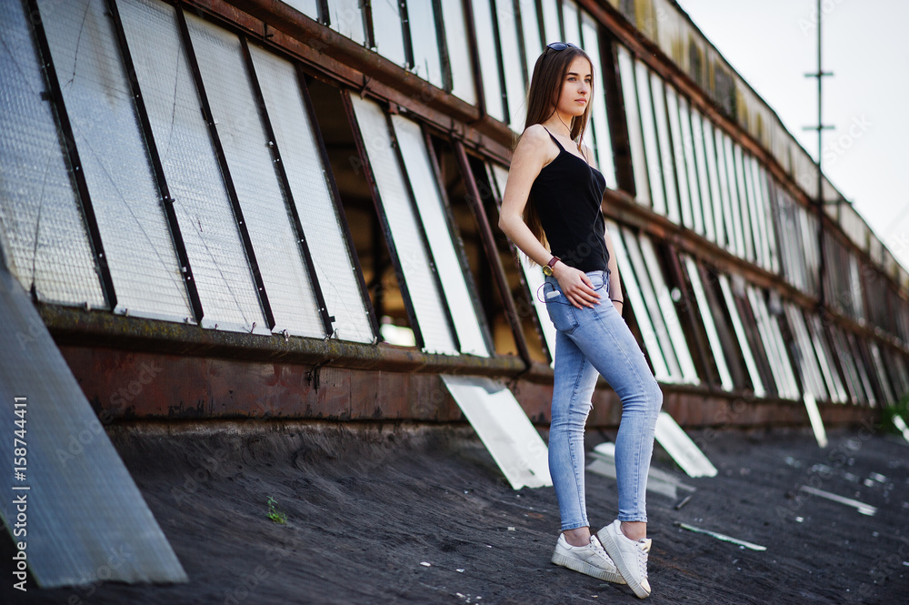 Girl at sunglasses and jeans, with phone and headphones posed at the roof of abadoned industrial place with windows.