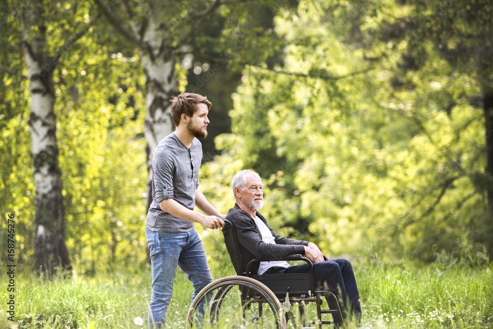 Hipster son walking with disabled father in wheelchair at park. Hipster son walking with disabled father in wheelchair at park.