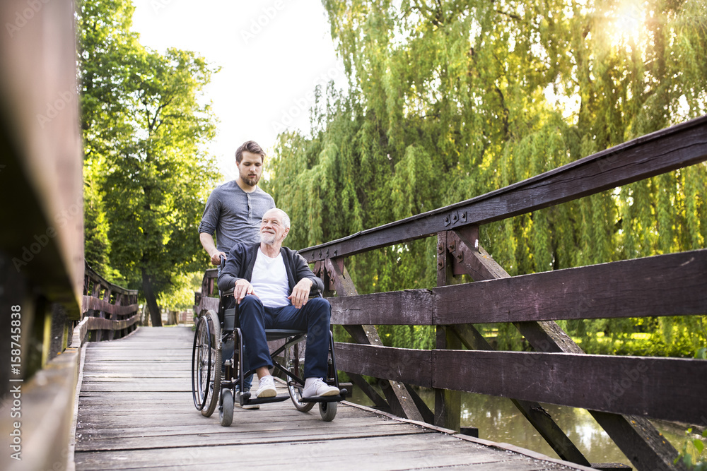 Hipster son walking with disabled father in wheelchair at park. Hipster son walking with disabled father in wheelchair at park.