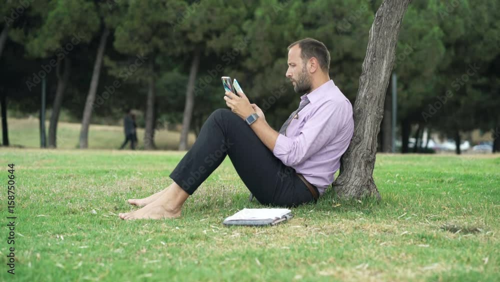 Young businessman working with smartphone and tablet sitting on grass in park Young businessman working with smartphone and tablet sitting on grass in park