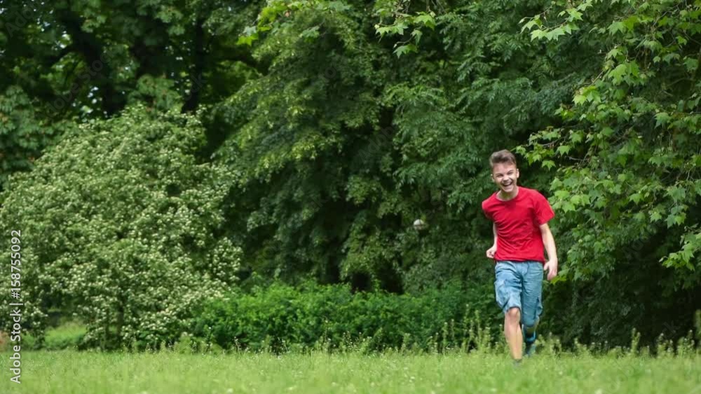 Happy teen boy running through the field towards the camera. Child running by in grass at summer park, slow motion. Happy teen boy running through the field towards the camera. Child running by in grass at summer park, slow motion.