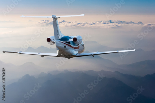 Private jet, passenger wide-body plane or aircraft is flying in the blue sky over the the clouds and mountains. Summer vacation concept