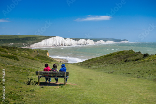 Three people sitting on a bench watching the waves on a windy but sunny day at Cuckmere Haven Sussex UK