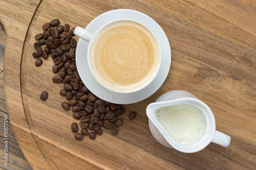 Coffee concept  Top view of  white porcelain cup filled with delicious frothy coffee  jug of milk and fresh roasted coffee beans on rustic wooden background.