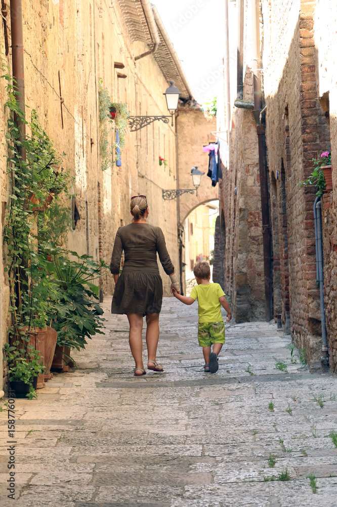 Woman and little boy are walking hand in hand through the narrow street of historical center of Colle di Val d'Elsa in Italy