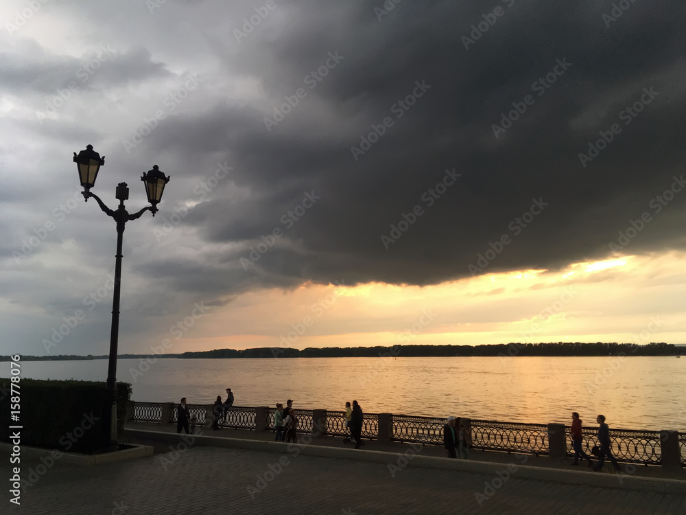 Темные дождевые тучи над набережной города и багровый закат над рекой

