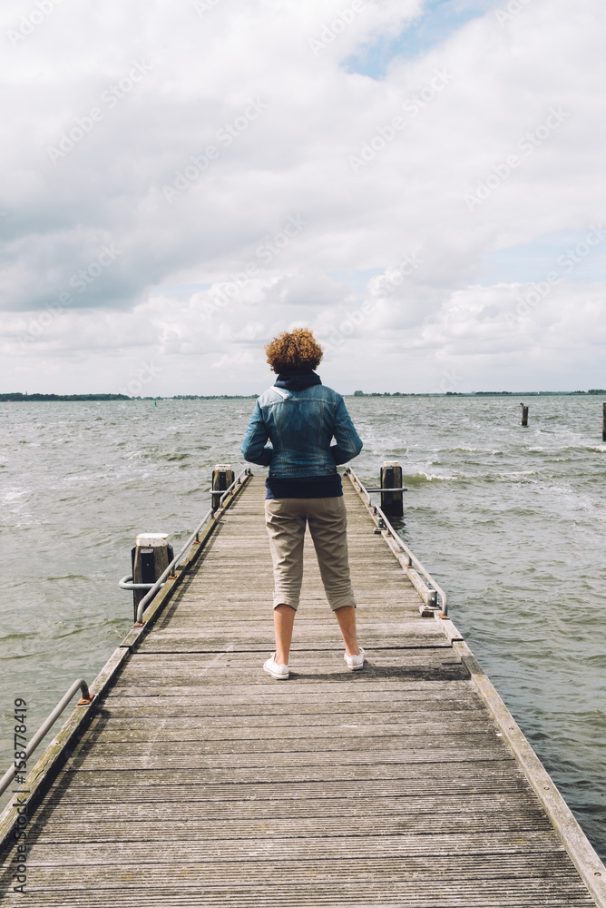 Back view of woman with casual clothing standing on wooden pier a cloudy summer day. Concept freedom Back view of woman with casual clothing standing on wooden pier a cloudy summer day. Concept freedom