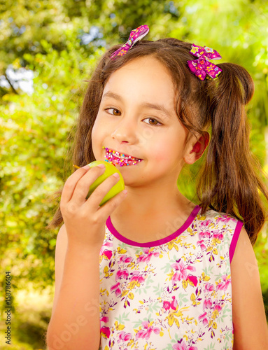 Beautiful young girl  eating a healthy apple and colorful dragees in her lips  in a blurred garden background