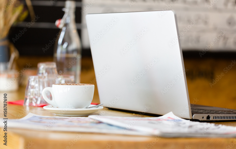 Silver open laptop, cup of coffee, newspaer and glass of water on wooden table in restaurant