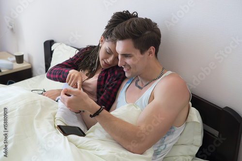 Young couple huggling in bed and looking at smartphone at home in the bedroom or hotel. Girl with zizi cornrows dreadlocks photo
