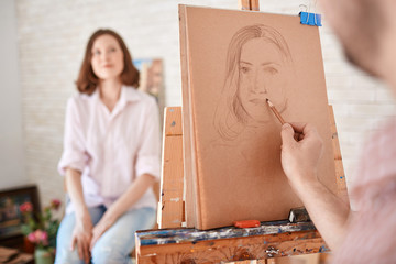 Portrait of artist drawing  portrait of young woman sitting in front of him in art studio, focus on the picture in pencil