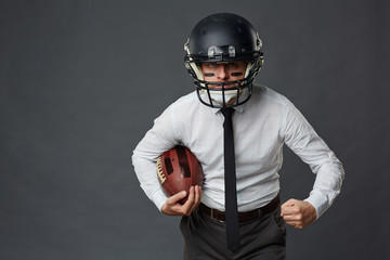 Aggressive middle-aged businessman with paint on his face wearing American football helmet and holding ball while looking at camera against grey background