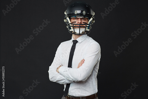 Waist up portrait of confident businessman wearing American football helmet standing cross-armed against black background and looking at camer