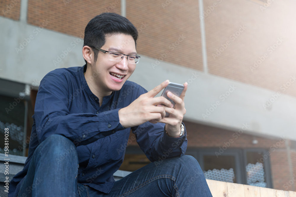 Asian man sitting and chatting by smartphone with modern building background. Asian man sitting and chatting by smartphone with modern building background.