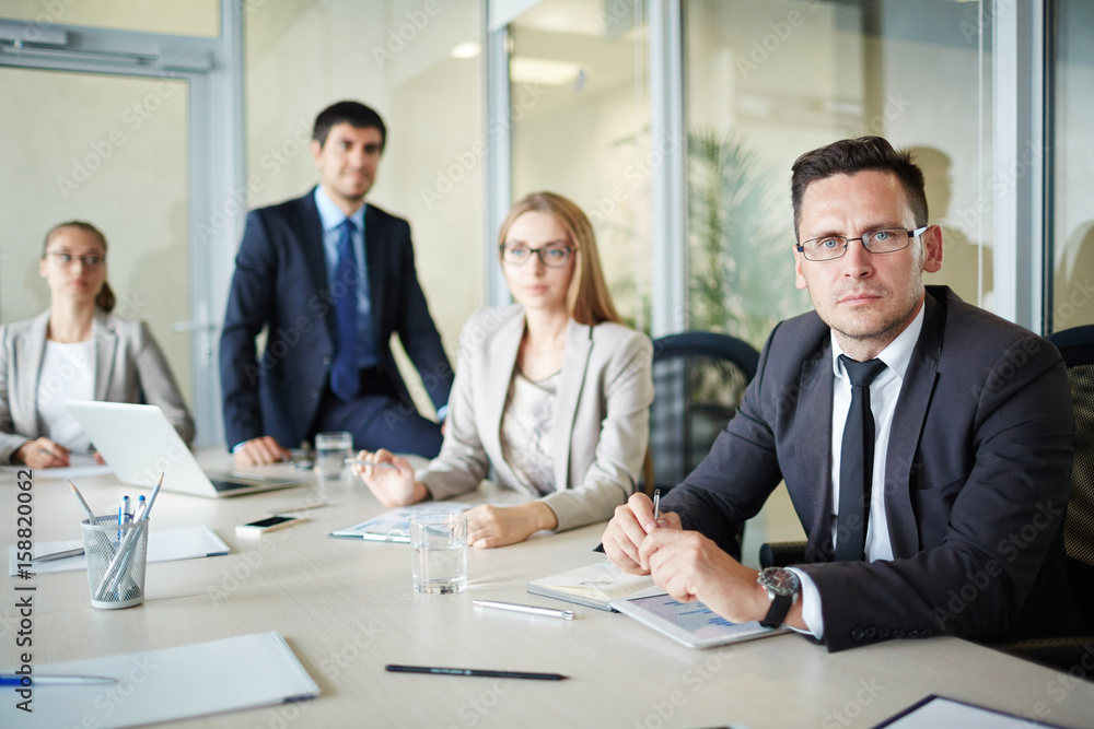 Waist-up portrait of serious middle-aged businessman in eyeglasses looking at camera with concentration while having board meeting Waist-up portrait of serious middle-aged businessman in eyeglasses looking at camera with concentration while having board meeting