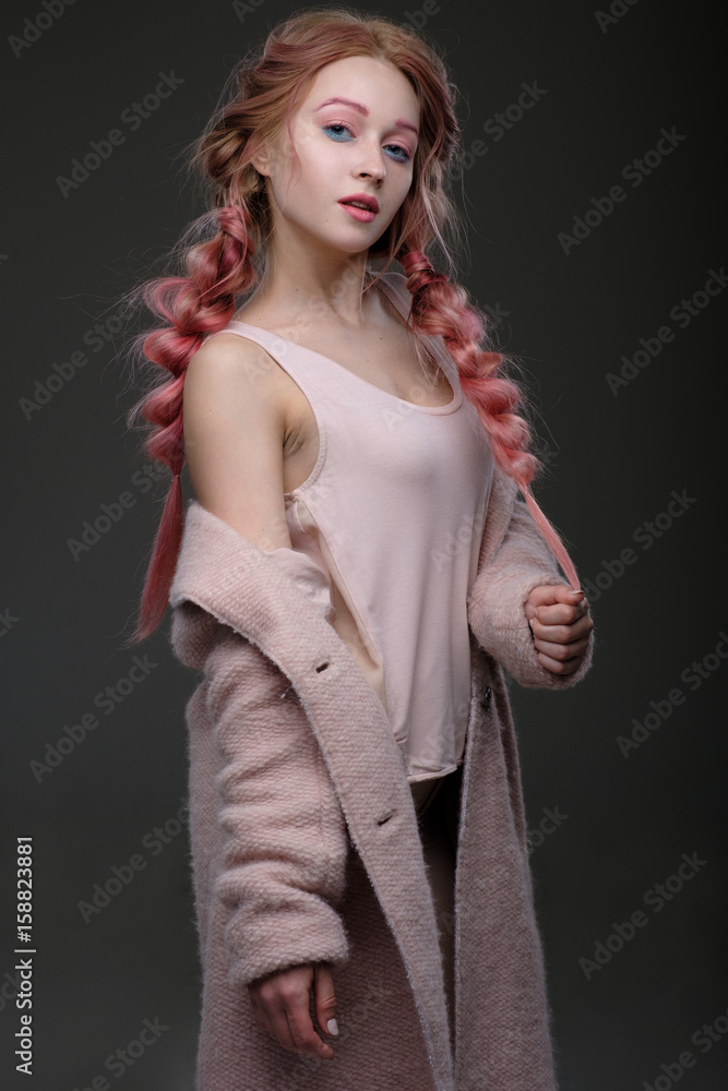 The girl with pink hair in braids, and pink-blue makeup with a pink coat worn off the shoulder stands in half-turn on a dark background, holding the tip of the braid and looking at the camera The girl with pink hair in braids, and pink-blue makeup with a pink coat worn off the shoulder stands in half-turn on a dark background, holding the tip of the braid and looking at the camera