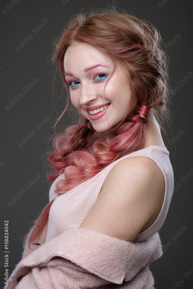 Fototapeta premium Close-up portrait of girl with pink hair in braids, and pink-blue makeup with a pink coat worn off the shoulder standing in the half-turn on a dark background smiling and looking at camera