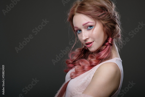 Close-up portrait of girl with pink hair in braids, pink and blue makeup, standing in a half-circle on a dark background playfully looking into the camera